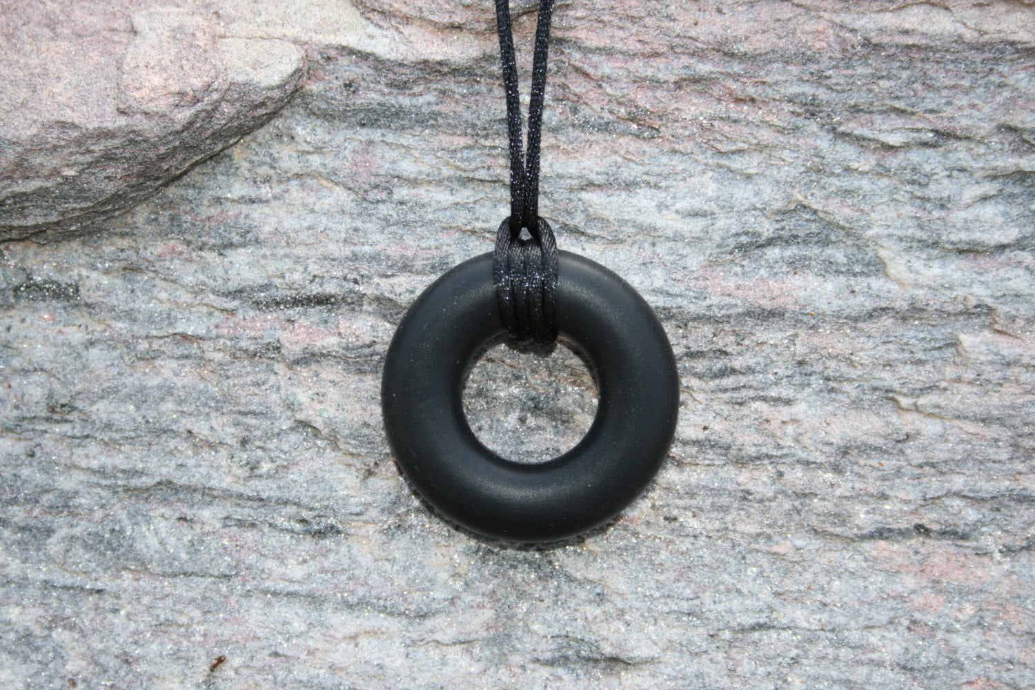 Pictured is black sensory Necklace for Children with Chewable Circle Pendant - available in 3 colors: black, turquoise, and navy blue! Measures Approximately 18" - This pendant and is made from food-grade silicone, strung on a nylon cord, and finished off with a plastic clasp designed to pop open when pulled really hard. (Jewelry on rock background)