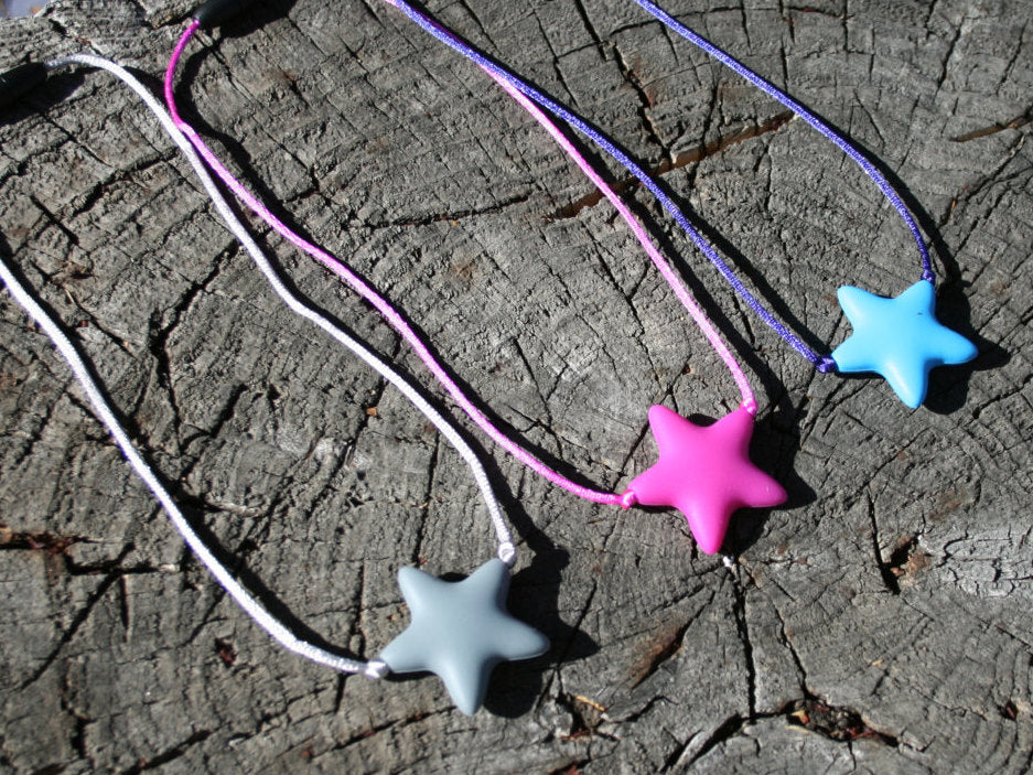 Sensory Necklace Chewable for Children with Star Pendant available in 3 colors: gray, hot pink, and aqua blue.  Measures approximately 18", but can be made longer if needed. This pendant and is made from food-grade silicone, strung on a nylon cord, and finished off with a plastic clasp designed to pop open when pulled really hard. (Jewelry on wooden background)