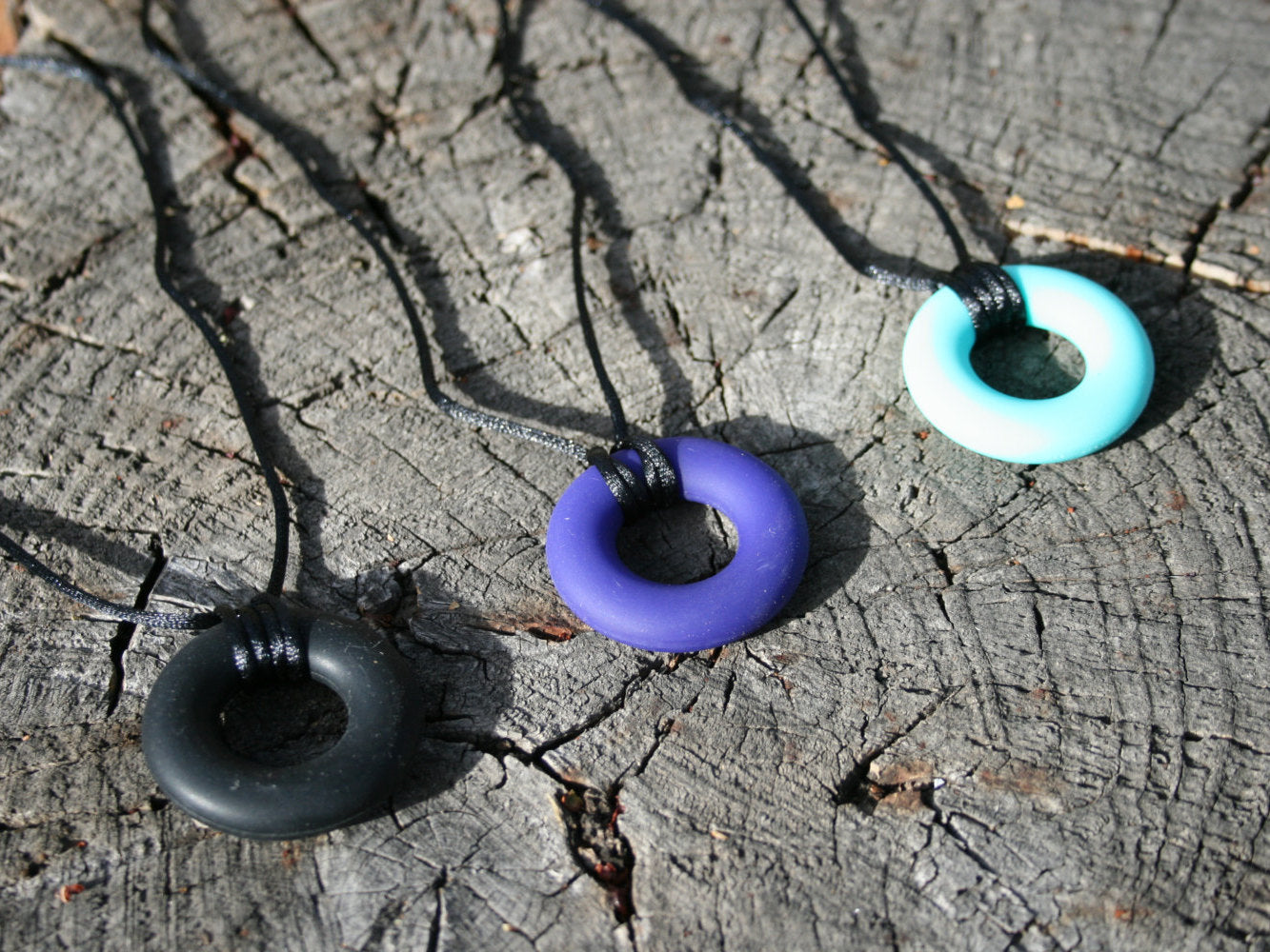 Pictured is variations of sensory Necklaces for Children with Chewable Circle Pendant - available in 3 colors: black, turquoise, and navy blue! Measures Approximately 18" - This pendant and is made from food-grade silicone, strung on a nylon cord, and finished off with a plastic clasp designed to pop open when pulled really hard. (Jewelry on wooden background)  
