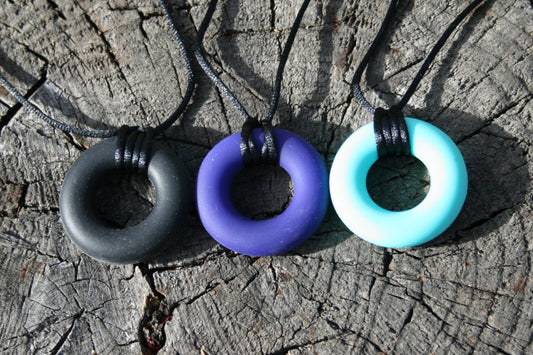 Pictured is variations of sensory Necklaces for Children with Chewable Circle Pendant - available in 3 colors: black, turquoise, and navy blue!  Measures Approximately 18" - This pendant and is made from food-grade silicone, strung on a nylon cord, and finished off with a plastic clasp designed to pop open when pulled really hard. (Jewelry on wooden background)