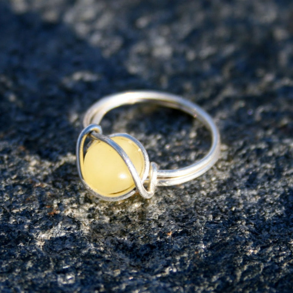 Picture of Baltic Amber Ring, Sterling Silver, Wire Wrapped, Unpolished, Raw, Any Size. This can be made with any color of amber: Milky(pictured), Lemon, Honey, Cognac, Coffee, or Green. The amber bead sits against the finger and allows the amber to work its magic! (Ring on rock background)