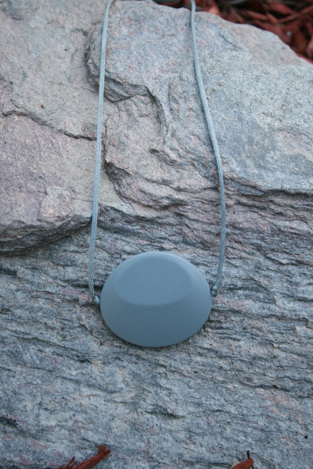 Picture of Chewable Sensory Necklace for Children with Large Oval Pendant and is made from food-grade silicone, strung on a nylon cord, and finished off with a plastic clasp. (Necklace on rock background)  
