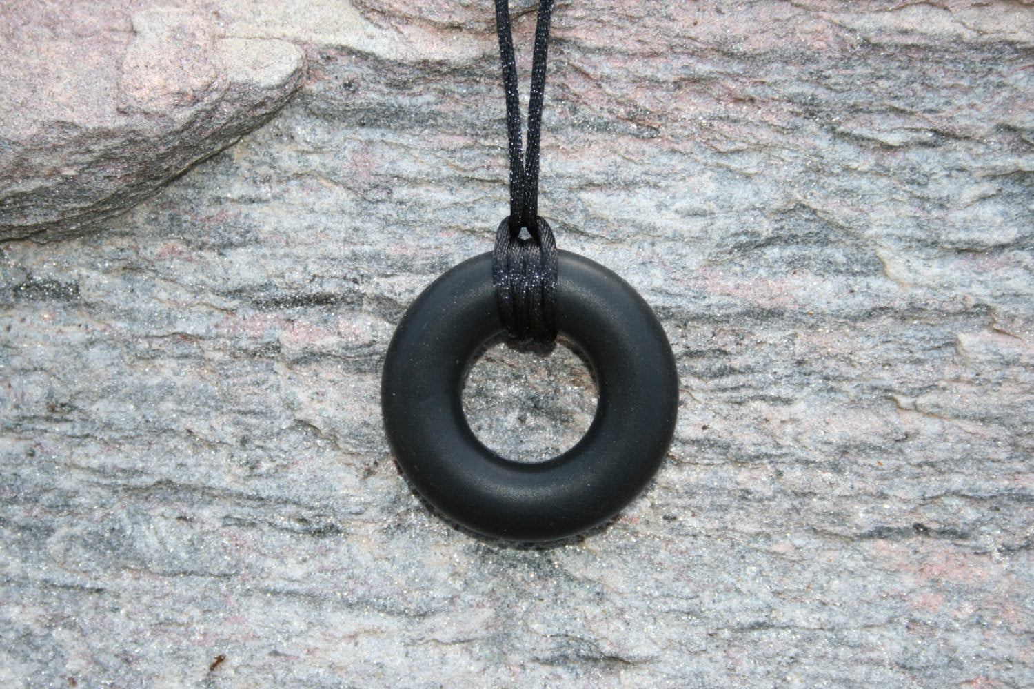 Pictured is black sensory Necklace for Children with Chewable Circle Pendant - available in 3 colors: black, turquoise, and navy blue! Measures Approximately 18" - This pendant and is made from food-grade silicone, strung on a nylon cord, and finished off with a plastic clasp designed to pop open when pulled really hard. (Jewelry on rock background)  